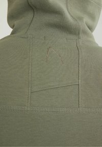 Felpa verde oliva con tessuto a costine, tasca a marsupio e un piccolo dettaglio del logo ricamato sulla schiena. Cuciture piatte visibili.