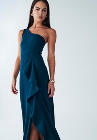 Vestido verde azulado de un solo hombro con corpiño ajustado y falda fluida con volantes, con una abertura alta y textura suave. Accesorio de pulsera dorada.