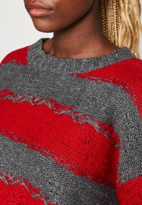 Närbild av en person som bär en röd och grå stickad tröja med slitna detaljer och rund halsringning.