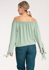 Jasnozielona bluzka typu off-the-shoulder z marszczonym dekoltem i wiązaniami na długich rękawach, zestawiona z ciemnoniebieskimi dżinsami.