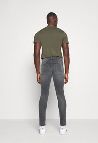 Muž stojící zády, oblečený v olivovém tričku, vybledlých šedých skinny džínách a bílých teniskách Adidas na jednobarevném pozadí.
