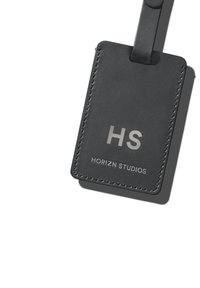 Schwarzes Ledergepäcketikett mit genähten Kanten, das silberne, geprägte Initialen "HS" und "HORIZN STUDIOS" deutlich zeigt.