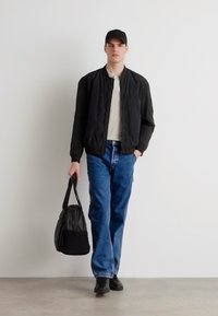 Chaqueta bomber acolchada negra sobre una camisa blanca, combinada con jeans de mezclilla azules. Gorra y zapatos negros. Sosteniendo una bolsa negra. Fondo neutro.