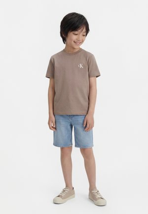 Αγόρι που φοράει κοντομάνικη μπλούζα σε χρώμα ταούπ με το λογότυπο "CK", ανοιχτόχρωμα τζιν σορτς και μπεζ αθλητικά παπούτσια, στέκεται και κοιτάζει προς τα κάτω.