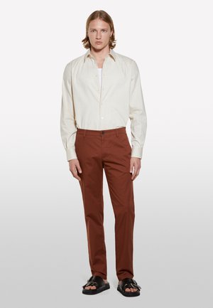 Chemise beige clair à boutons, à manches longues, associée à un pantalon couleur rouille. Des sandales noires complètent la tenue. Design simple et ajusté.