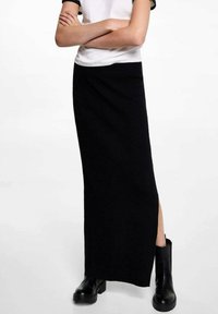 Černá pletená maxi sukně s bočním rozparkem, v kombinaci s bílým a černým topem s krátkým rukávem. Modelka má na sobě černé kotníkové boty.