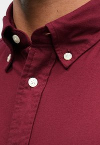 Bordo košulja s dugim rukavima od teksturirane tkanine, s izrezom u obliku koplja, dva bijela gumba i urednom vrstom detalja.