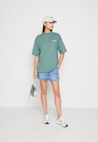 Oversized teal T-shirt with logo detail, paired with a denim mini skirt and white sneakers. Accessory: small iridescent handbag and white cap.