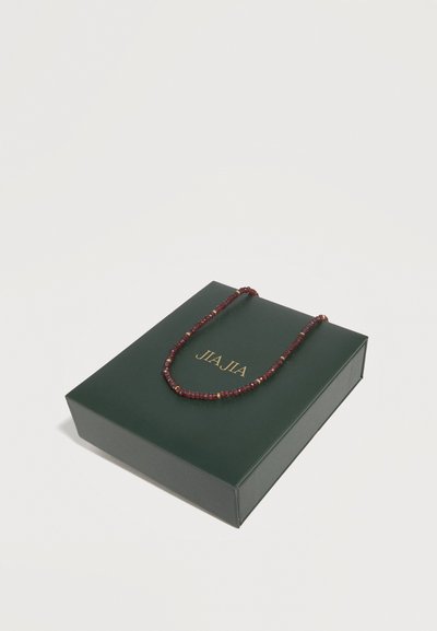 Σκούρο πράσινο κουτί με χρυσό κείμενο "JIA JIA" και ένα κόκκινο κολιέ με χάντρες τοποθετημένο από πάνω, σε απλό λευκό φόντο.