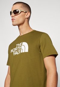 Grønn bomulls t-skjorte med et hvitt logodesign på forsiden. Modell med beige sports solbriller med brune linser, vendt mot venstre.