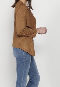 Femme portant une veste en daim marron clair et un jean bleu, vue de profil sur un fond blanc uni.