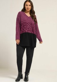 Femme portant un pull coloré en noir et violet avec imprimé léopard, un pantalon noir et des bottines noires se tenant devant un fond neutre.