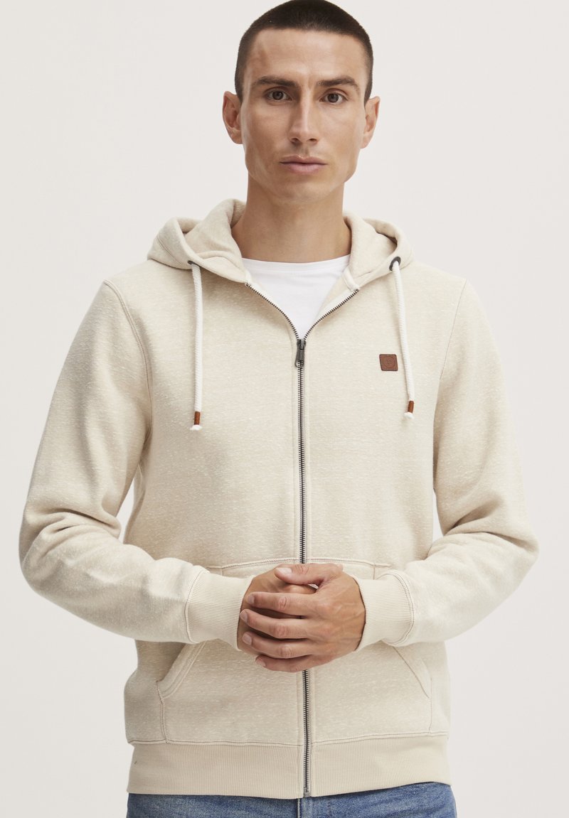 Lys beige zip-hette med teksturert overflate, justerbar hette, frontlommer og en liten logo-lapp på venstre bryst.