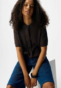 Vrouw met krullend haar, gekleed in een zwart knoopshirt met korte mouwen en blauwe spijkerbroeken, zittend met haar handen tussen haar benen.