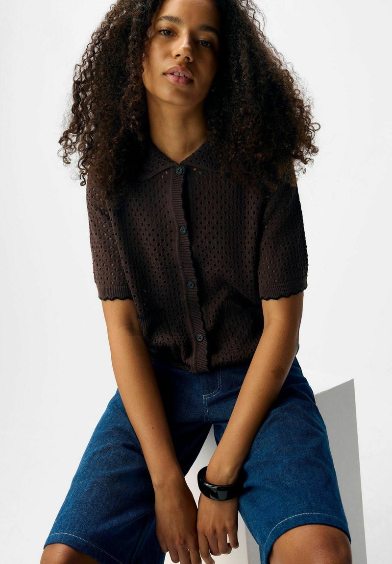 Kobieta z kręconymi włosami, ubrana w czarną koszulę z krótkim rękawem zapinaną na guziki i niebieskie dżinsowe szorty, siedzi z rękami opartymi na udach.