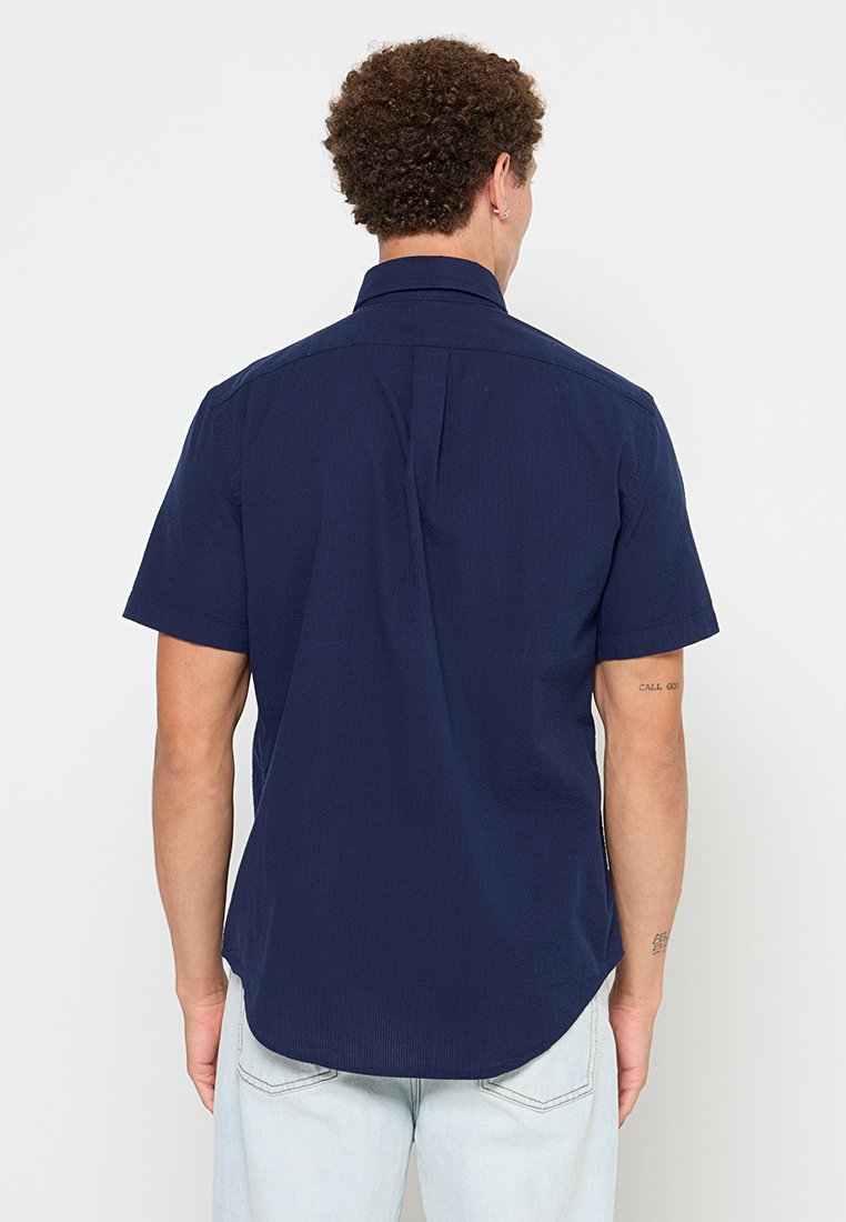 Chemise à manches courtes bleu marine avec un tissu légèrement texturé, un col classique et un ourlet droit. Présente un pli central au dos.