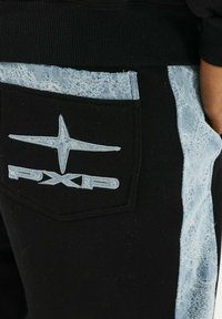Pantalones de chándal negros con acentos de denim azul claro. Presentan un bolsillo trasero con un logotipo en bordado blanco y un patrón texturizado similar al denim.