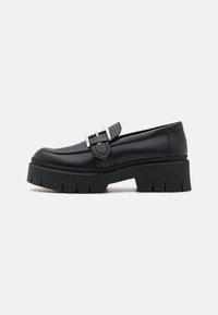 Mocassins en cuir noir avec un bout arrondi, présentant un détail de boucle sur le côté et une semelle plateforme épaisse avec des rainures prononcées.