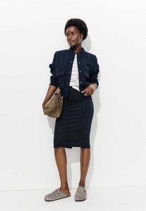 Femme portant une veste bleu marine, une jupe noire jusqu'aux genoux, une chemise rayée, des chaussures à enfiler à imprimé léopard, et tenant un sac à main en daim marron, se tenant devant un mur blanc.