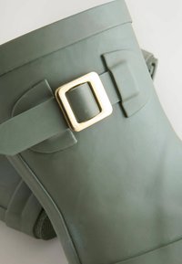 Stivale di gomma verde scuro con un occhiello in oro metallizzato e una superficie piatta e testurizzata progettata per la durata.