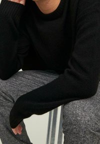 Pull en maille noire à manches longues avec un col rond côtelé. Pantalon gris à motifs visible, mettant en valeur le contraste des textures.