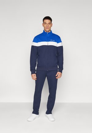 Navy tracksuit featuring a blue and white color-block design, made from soft fabric, with a zip-up jacket and tapered pants. White sneakers.