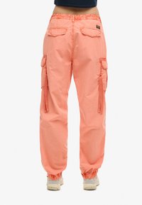 Persoon in koraalkleurige cargo broek met elastische boorden en zijzakken, staande op blote voeten tegen een effen witte achtergrond.