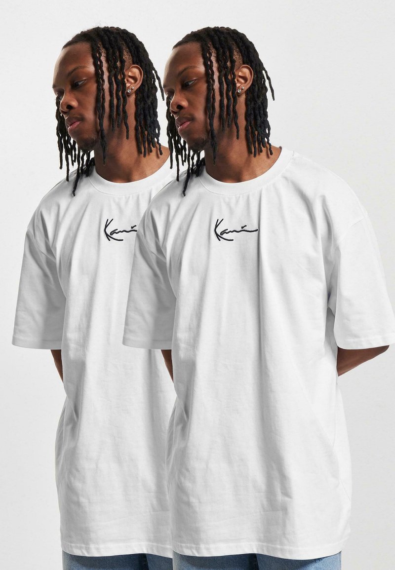 White oversized cotton t-shirt featuring a black embroidered logo on the chest. Model shown from the side with loose-fitting sleeves.