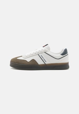 Zapatilla blanca y marrón con puntera de ante texturizado, cuerpo de cuero suave y suela de goma oscura. Presenta acentos en azul y rojo, y una marca.