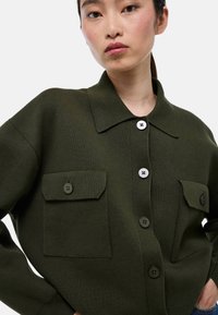 Gilet en tricot vert olive avec un col, deux poches avant et de grands boutons. Le tissu texturé ajoute de la profondeur.