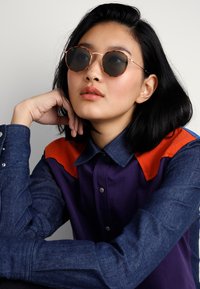 Ochelari de soare rotunzi cu lentile colorate, hardware auriu și un cadru cu un model. Modelul poartă o cămașă din denim albastru cu un design portocaliu și mov.