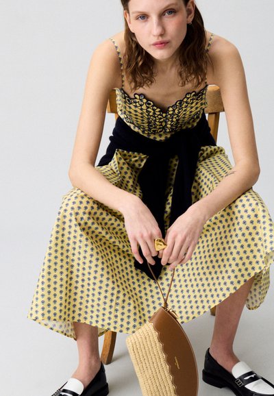 Abito floreale giallo con accenti blu navy, spalline sottili e gonna a pieghe, abbinato a un maglione nero legato in vita e scarpe nere.