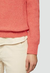 Korallfarbener Strickpullover mit gerippter Textur und runder Saum, getragen über einem cremefarbenen, geschichteten Hemd und hellen Hosen.