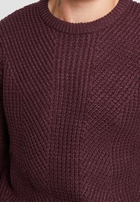 Primo piano di un maglione lavorato a maglia bordeaux con un motivo a coste diagonali texturizzato e un collo rotondo.