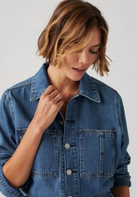 Veste en denim avec un col, fermeture à boutons et deux poches à l'avant. Présente une couleur bleu clair et une texture légèrement délavée.