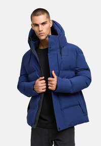 Mladý muž, ktorý si upravuje zips na modrej prešívanej zimnej bunde s kapucňou, oblečený v čiernom tričku a tmavých nohaviciach, na jednofarebnom pozadí.