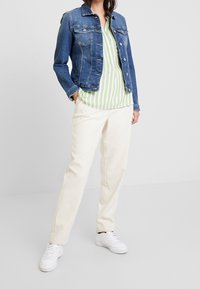 Chaqueta de mezclilla con dos bolsillos en el pecho, combinada con una blusa a rayas verde y blanca, pantalones crema y zapatillas blancas. Ajuste casual.