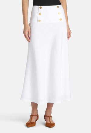 Femme portant une jupe midi blanche taille haute avec six boutons dorés et des chaussures à bride en T pointues marron, debout sur un fond blanc.