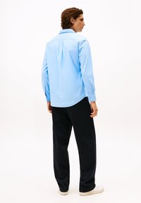 Hellblaues Hemd mit langen Ärmeln, Rückenschlitzdetail und abgerundetem Saum, kombiniert mit dunklen, locker sitzenden Hosen und weißen Schuhen.
