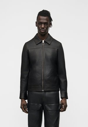 Mladý muž s tmavou pokožkou a krátkymi dredmi, oblečený v čiernej koženej bunde na zips a zladených čiernych kožených nohaviciach s nákladnými vreckami.