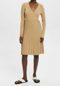 Stickad, beige omslagsklänning med långa ärmar, plisserad kjol och V-ringning. Kombinerad med vita sandaler med svart sula.