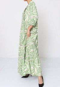 Robe maxi verte à volants avec motif de feuilles et manches à volants, ornée de dentelle et à la silhouette fluide. Portée avec des talons aiguilles noirs.