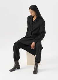 Costum negru, croit cu sacou și pantaloni conici, asortat cu botine maro. Textură netedă, design minimalist, fără modele vizibile.