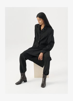 Costume noir ajusté avec un blazer et un pantalon fuselé, associé à des bottines marron. Texture lisse, design minimaliste, pas de motifs visibles.