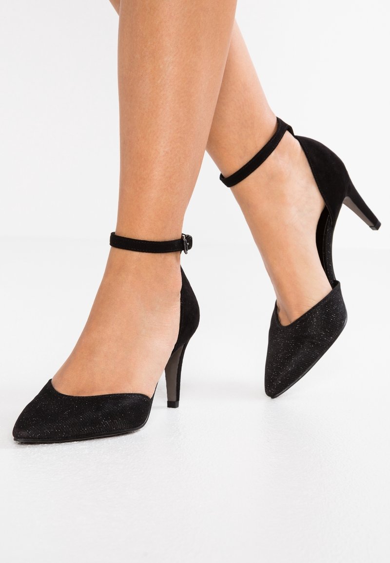 Pieds portant des talons hauts noirs à bout pointu avec des brides à la cheville, un pied à plat et l'autre légèrement levé.