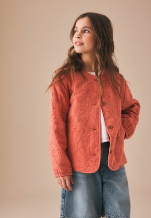 Ung pige med langt brunt hår iført en tekstureret orange knapjakke og denimbukser, der kigger mod venstre mod en beige baggrund.