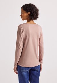 Femme aux cheveux bouclés portant une chemise à manches longues rose clair et un jean bleu, se tenant de dos devant un fond blanc.