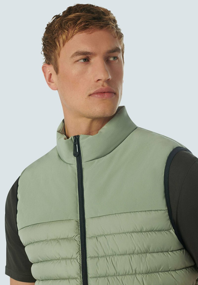 Gilet rembourré vert clair avec un col haut, fermeture éclair sur le devant et manches sombres contrastantes. Il présente une texture matelassée et un tissu lisse.