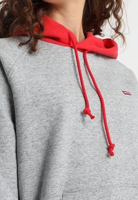 Sweat à capuche gris avec une doublure intérieure rouge et des cordons, présentant un petit logo sur la poitrine, et un tissu doux et texturé.