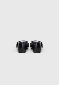 Sapatilhas planas de couro preto com ponta arredondada e uma fita fina no tornozelo, decorada com um laço na parte de trás. Textura suave e design minimalista.
